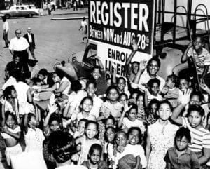 A group of African American children gather around a sign and booth to register voters in the early 1960s. PHOTO: FREEPIK/WIKIPEDIA